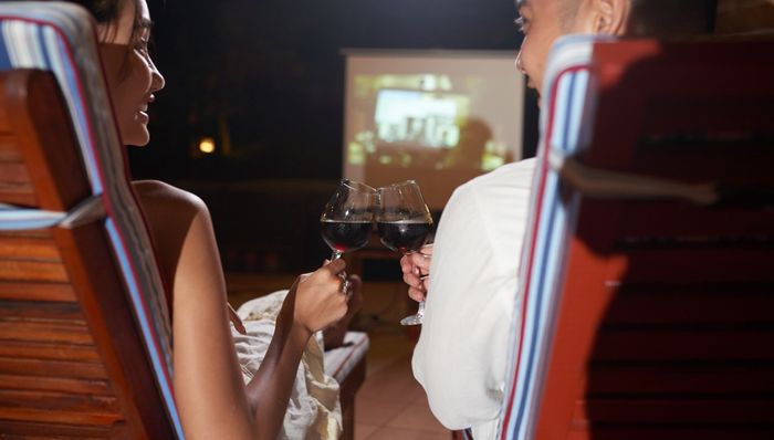 A couple toast with wine glasses as they watch a movie outside on a projector screen.